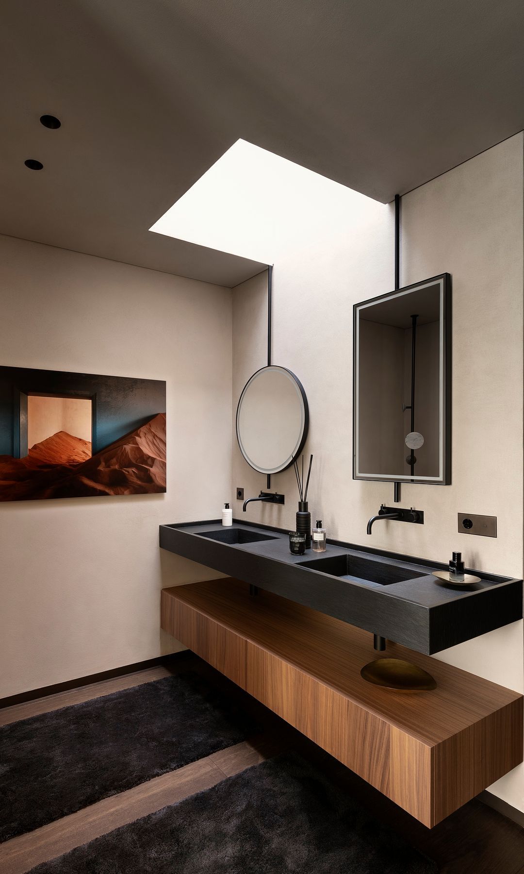 Baño con fregadero negro doble, muebles de madera, piso de madera, ventana del techo, espejo redondo y espejo rectangular
